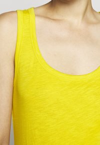 Keltainen hihaton paita on valmistettu pehmeästä ja joustavasta kankaasta. Siinä on avara kaula-aukko ja leveät olkaimet. Pintatekstuurissa on sileä vaikutelma, jossa on pieniä vaihteluita.