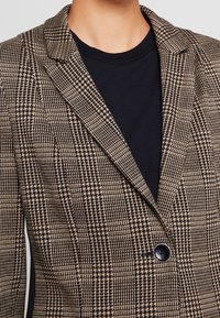 Blazer à motifs marron et noir avec un revers pointu, une fermeture à un bouton, et un tissu texturé. Porté sur une chemise noire.