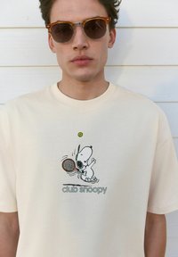 Camiseta beige con un gráfico de Snoopy jugando al tenis, con el texto verde "club snoopy" debajo de la ilustración, mangas cortas y textura suave.