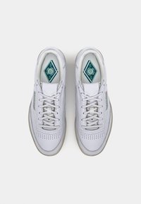 Λευκά δερμάτινα αθλητικά παπούτσια με στρογγυλεμένες μύτες, διάτρητες λεπτομέρειες και γκρι ελαστικές σόλες. Πράσινες λεπτομέρειες στις εσωτερικές σόλες και τη γλώσσα.