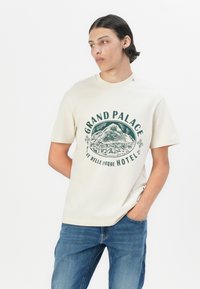 Beige t-skjorte med korte ermer som har grønn grafisk tekst og fjellillustrasjon. Laget av bomull med avslappet passform, rund hals og uten dekorasjoner.