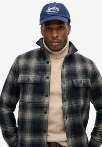 Mørkeblå kasket med hvidt logo, ternet flannel skjorte i navy og creme, og en beige turtleneck. Skjorten har klaplommer og knaplukning.