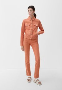 Pomarańczowa dżinsowa kurtka z długimi rękawami, z zapięciem na guziki z przodu, z dwoma kieszeniami na piersi, zestawiona z dopasowanymi pomarańczowymi dżinsami i jasnymi sandałami.