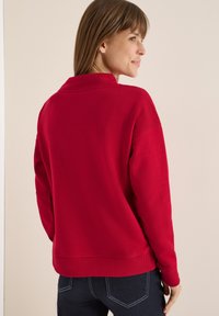 Czerwony sweter z wysokim kołnierzem i ściągaczami na mankietach, pokazany od tyłu. Tkanina wydaje się miękka i ma luźny krój. Widoczne granatowe dżinsy.
