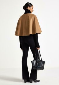 Tan uldekappe over sort højhalset trøje, sorte bredbenede bukser, holder en struktureret sort håndtaske. Glat tekstur, kappe hænger elegant.