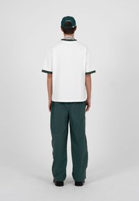 Person, der står med ryggen til og har en hvid kortærmet skjorte med mørk kant, mørkegrønne bukser, sorte sko og en grøn baseballkasket på.