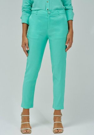 Mujer vestida con pantalones tobilleros de talle alto turquesa y camisa a juego, combinados con tacones de tiras beige, de pie contra un fondo liso.
