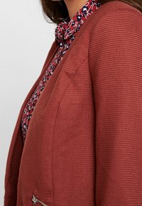 Ribbed rust-colored jacket featuring a zippered pocket, worn over a floral-patterned navy and red shirt with a buttoned collar.