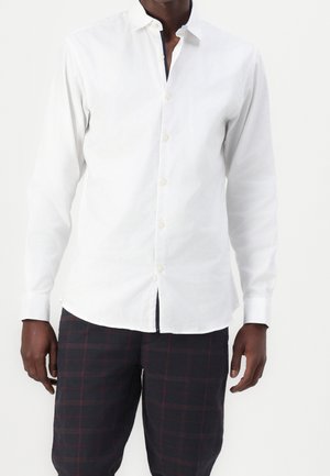 Man wearing a long-sleeve white button-up shirt and dark plaid trousers, standing against a plain light background.