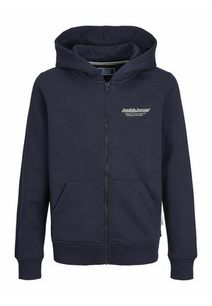 Barn iført navyblå hættejakke med lynlås og frontlommer, mørkeblå jeans og hvide sneakers, stående mod en ensartet lys baggrund.