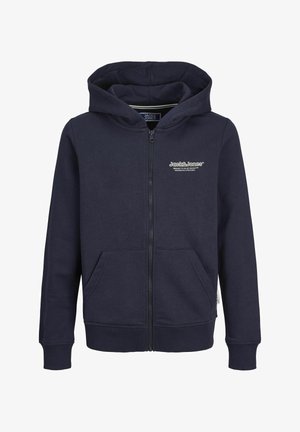Kind trägt marineblauen Zip-up-Hoodie mit Fronttaschen, dunkelblaue Jeans und weiße Turnschuhe und steht vor einem schlichten hellen Hintergrund.