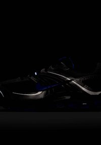 Svart sportskor med texturerad ovandel, blå accenter och reflekterande detaljer. Böjd design med vaddering synlig i sulan.