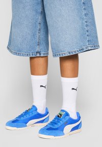 Blå mockaskor med vita detaljer, snörningsdesign och grön logotyp. Bärs med vita ribbade strumpor som har en svart logotyp. Jeansshorts.