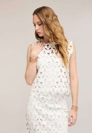 Jeune femme aux longs cheveux ondulés portant une robe blanche sans manches avec des motifs en broderie anglaise, regardant vers le bas, avec des bracelets dorés aux deux poignets.