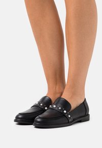 Black leather loafers with a rounded toe, featuring a wide strap adorned with white pearls and silver studs. Flat sole, minimalist design.
