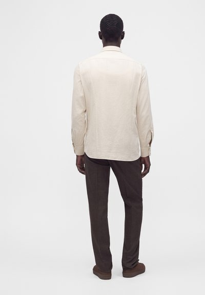 Homme debout, de dos, portant une chemise crème à manches longues, un pantalon marron foncé et des chaussures marron sur un fond blanc uni.