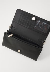 Portefeuille noir en cuir lisse et tissu texturé, avec une poche zippée, des emplacements pour cartes et une fenêtre ID transparente, sécurisé par un bouton pression.