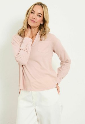 Rosa Langarmshirt mit V-Ausschnitt und aufgesetzter Tasche vorne, aus weichem Stoff gefertigt. Verfügt über geknöpfte Manschetten und eine bequeme Passform, kombiniert mit weißen Hosen.