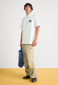 Γκρι κοντομάνικο πουκάμισο με ψηλό γιακά και κεντημένο λογότυπο, συνδυ ασμένο με χαλαρές μπεζ παντελόνες και τσάντα από μπλε τζιν. Μαύρα παπούτσια.