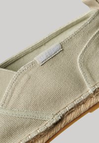 Zapato de lona color beige claro tipo slip-on con detalles de yute, bordes cosidos y una parte superior texturizada. Presenta una sutil etiqueta de "Superdry".