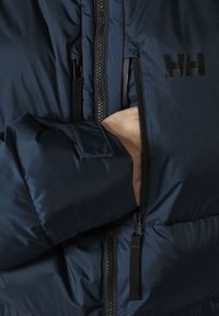 Chaqueta acolchada azul marino con un bolsillo con cremallera. Presenta una tela suave, textura acolchada y un logotipo en el pecho. El detalle de la cremallera es visible.