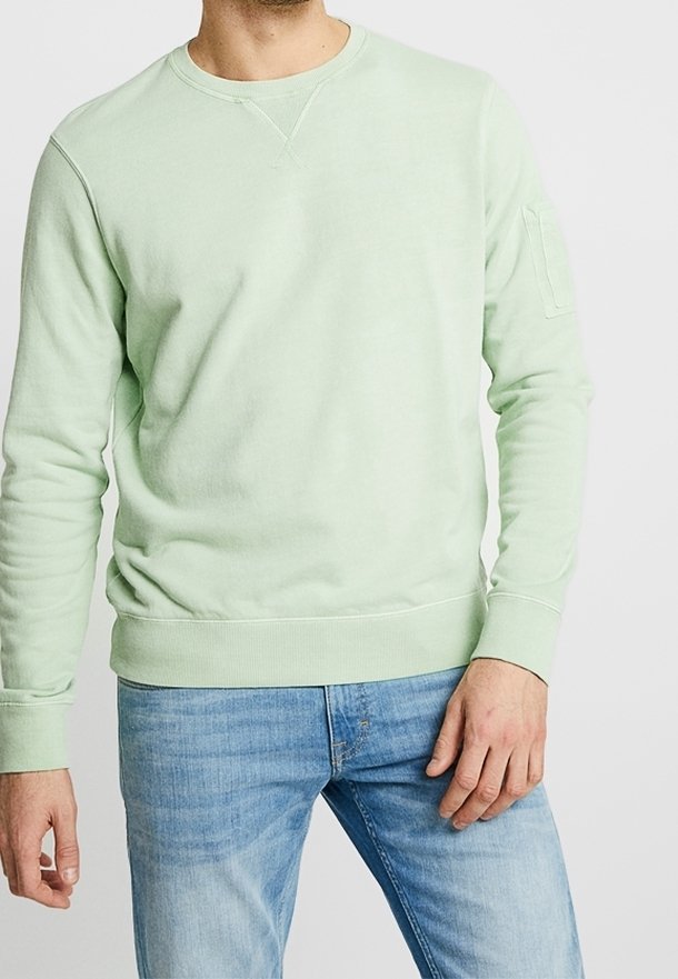 Sudadera verde menta de algodón suave, con cuello redondo, puños y dobladillo acanalados, que presenta un bolsillo en el pecho. Combinada con vaqueros azul claro.