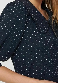 Námořnická modrá blůza s bílými puntíky, s kulatým límcem a krátkými nažehlenými rukávy. Měkká tkanina s hladkým povrchem a jemným leskem.