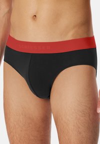 Sort boksershorts med rød elastisk talje, der har mærkenavnet "SCHIESSER" præget. Glat stof med en tætsiddende pasform.