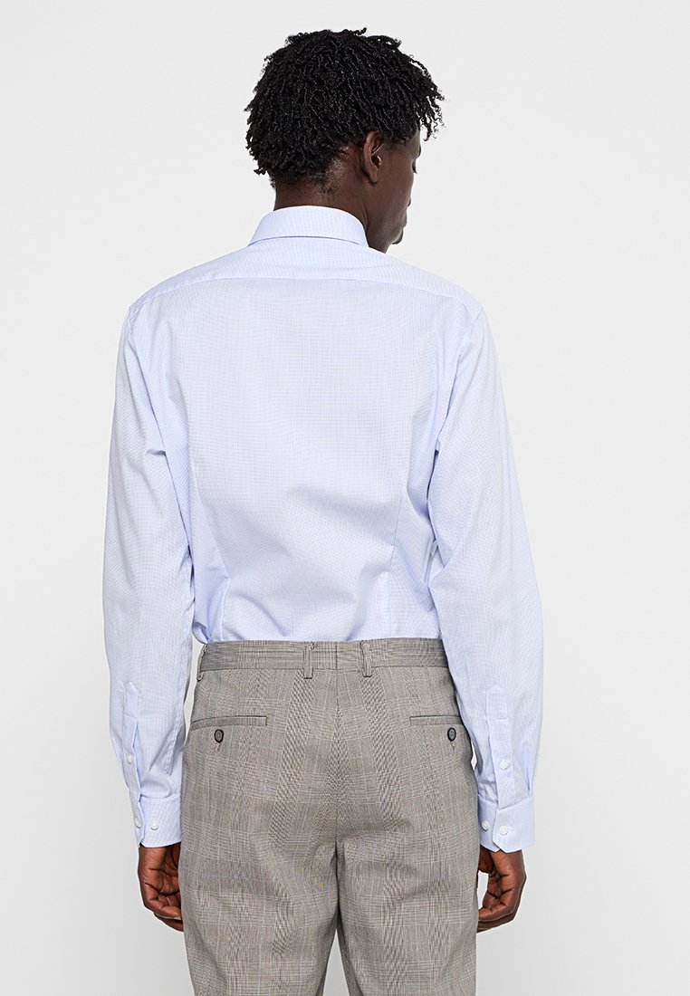 Hombre con cabello corto y rizado, vestido con una camisa de vestir de manga larga color azul claro y pantalones de cuadros grises, de pie de espaldas contra un fondo blanco.