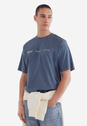 T-shirt en coton bleu à manches courtes, col rond et motifs textuels dans différentes polices. Associé à un denim bleu clair, avec un pull crème noué à la taille.