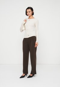 Long-sleeve cream top with a curved button placket, paired with high-waisted brown trousers and black pointed-toe heels. Holds a small bag.