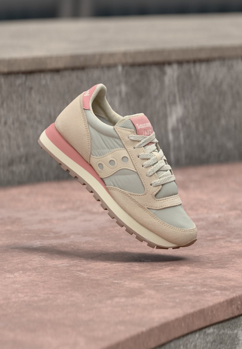 Ανοιχτό μπεζ και ροζ αθλητικό παπούτσι Saucony Jazz με λευκά κορδόνια, ενδιάμεση σόλα και υφασμένη λαστιχένια εξωτερική σόλα, να αιωρείται πάνω από μια πέτρινη επιφάνεια.