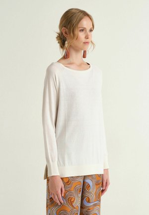 Frau mit blondem Haar im niedrigen Dutt trägt ein weißes Langarmoberteil, große orangefarbene Quastenohrringe und gemusterte orange-blaue Hosen, blickt nach rechts.