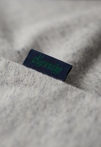 Grå stof med en blød tekstur har et marineblåt mærke med teksten "Superdry" i grøn syning, delvist stukket ind i materialet.