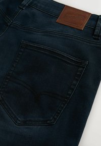 Tumed sinise teksapüksid, millel on tagatasku, mis on kaunistatud õrna kaarekujulise tikkimismustriga, ning selle kohal pruun nahkümbris.