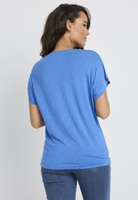 Niebieska bluzka z krótkim rękawem o luźnym fasonie, z małymi, wielokolorowymi kropkami. Wykonana z miękkiego materiału, ma okrągły dekolt i zaokrąglony dół.