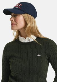Marineblaue Baseballmütze mit besticktem Logo, kombiniert mit einem grünen Strickpullover mit Kabelmuster, der ein kleines Logo auf der Brust und einen weißen Kragen hat.
