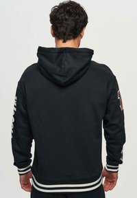 Felpa nera con cappuccio regolabile, polsini e orlo a coste. Presenta strisce bianche e una grafica rossa sulla manica sinistra. Texture morbida.