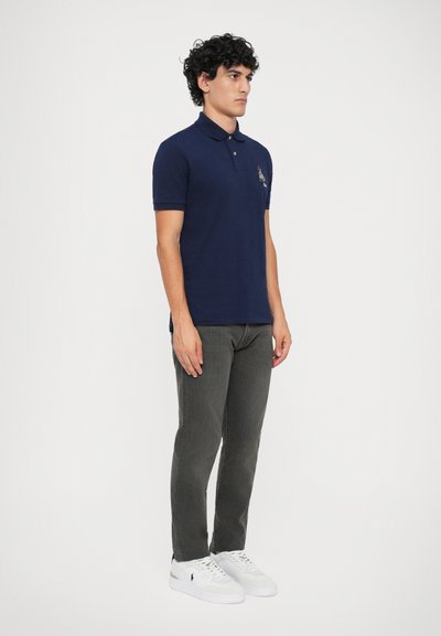 Camisa polo azul-marinho com colarinho, mangas curtas e logótipo bordado. Usada com calças cinzentas e sapatilhas brancas, mostrando um estilo casual.