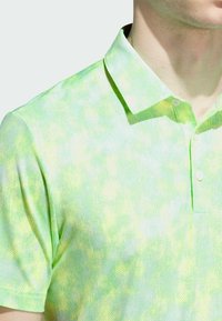 Zielona koszulka polo z fakturowanej tkaniny, z subtelnym wzorem tie-dye w jaśniejszych odcieniach. Kołnierz jest płaski, zapinany na dwa guziki.