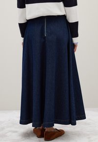 Personne portant une longue jupe en denim bleu foncé avec une fermeture éclair arrière visible, des chaussures plates marron et un haut rayé noir et blanc.