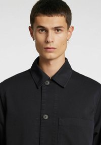 Mustast kangast nööbitav jakk, terava krae ja rinnataskuga, mati tekstuuri ja kahe nööbiga ees.