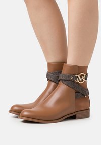 Bottines en cuir marron avec une bride en tissu foncé, ornées d'une quincaillerie dorée et d'un accent logo, bout rond et talon bloc bas.