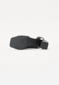 Musta korkokenkä, jossa on leveä nauha ja avonaisella varvasosalla. Varustettu teksturoidulla kumipohjalla ja neliömäisellä korolla. Minimalistinen muotoilu.