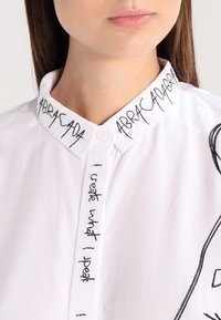 Camicia bianca con bottone e testo ricamato "ABRACADABRA" sul colletto e "Creo ciò che dico" sulla parte anteriore; texture liscia.