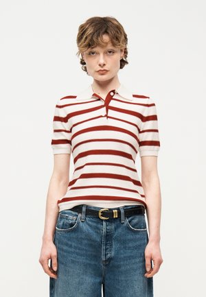 Rövid, göndör hajú személy fehér és piros csíkos, rövid ujjú galléros pólót visel, amely kék farmernadrágba van betűrve, kiegészítve egy fekete övvel.