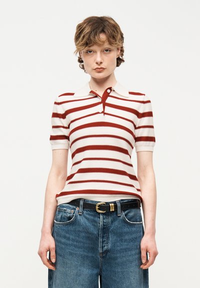 Personne aux cheveux courts et bouclés portant un polo à manches courtes blanc et rouge rayé, rentré dans un jean bleu avec une ceinture noire.