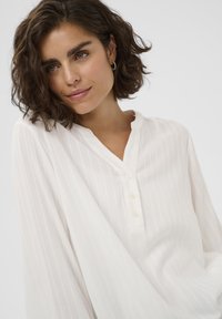 Femme avec des cheveux courts et bouclés portant une blouse blanche à rayures verticales et un col boutonné, regardant légèrement vers le bas avec un doux sourire.