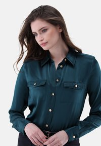 Blusa di seta turchese con colletto, bottoni dorati e due tasche frontali. Tessuto liscio e maniche lunghe con polsini abbottonati.