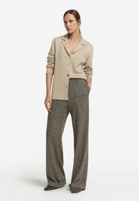 Beige knäppbar cardigan med långa ärmar, parat med lösa, texturerade, mönstrade bruna byxor. Modellen står med händerna i fickorna.